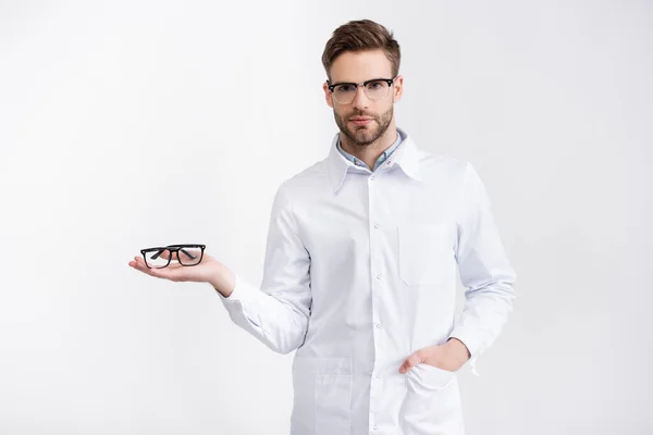 Vue de face du médecin confiant avec la main dans la poche tenant des lunettes sur la paume, tout en regardant la caméra isolée sur blanc — Photo de stock