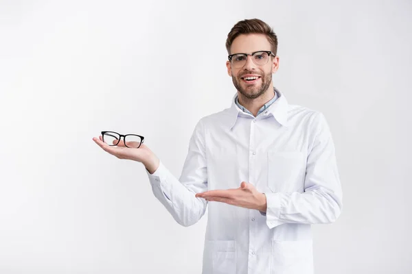 Vista frontal del médico feliz sosteniendo y apuntando con la mano a las gafas en la palma, mientras mira a la cámara aislada en blanco - foto de stock