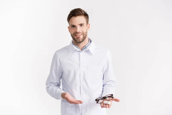 Front view of smiling doctor pointing with hand at eyeglasses and lenses container on palm isolated on white — Stock Photo