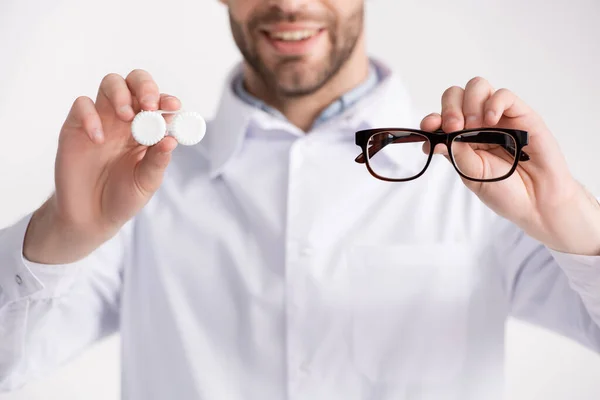 Ausgeschnittene Ansicht eines lächelnden Arztes mit Händen in der Luft, zeigt Brille und Linsenbehälter isoliert auf weißem Hintergrund auf verschwommenem Hintergrund — Stockfoto