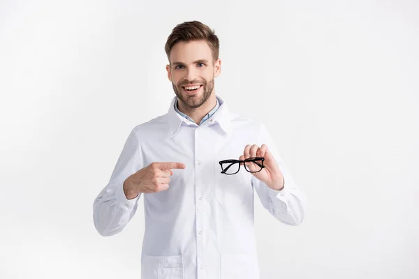 Vue de face du médecin heureux pointant du doigt les lunettes, tout en regardant la caméra isolée sur blanc — Photo de stock