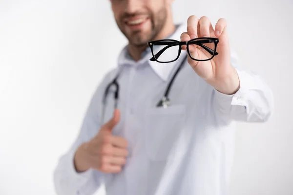 Vue recadrée du médecin le pouce en l'air, montrant des lunettes à monture noire isolées sur fond blanc sur fond flou — Photo de stock