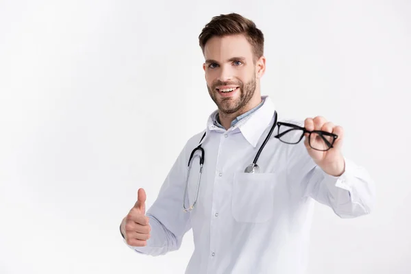 Smiling ophthalmologist with stethoscope, showing approval gesture, while showing eyeglasses on blurred foreground isolated on white — Stock Photo