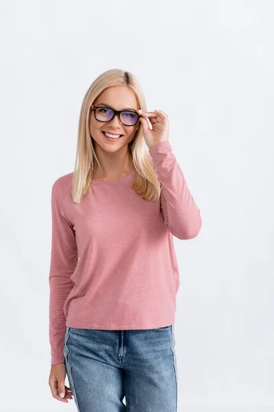 Smiling blonde woman in jeans and pink long sleeve holding eyeglasses frame, while looking at camera isolated on white — Stock Photo