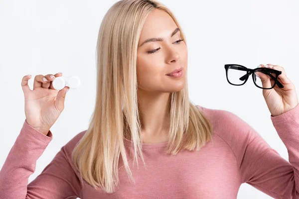 Blonde woman looking at eyeglasses while holding contact lenses isolated on white — Stock Photo