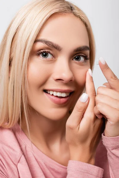 Blonde Frau lächelt, während sie Kontaktlinse trägt und die Wange berührt, isoliert auf grau — Stockfoto