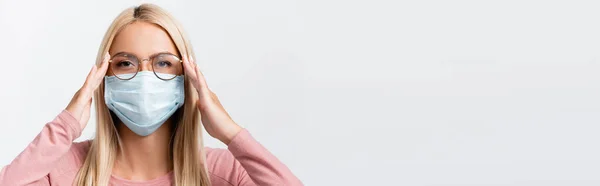Femme blonde en masque médical touchant les lunettes isolées sur gris, bannière — Photo de stock