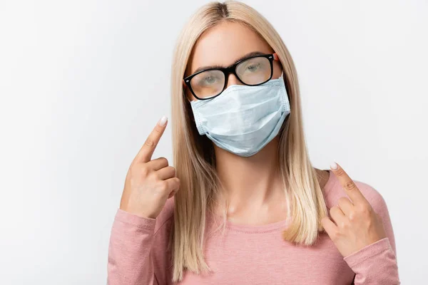 Mujer joven con máscara médica apuntando con el dedo a las gafas empañadas aisladas en gris - foto de stock