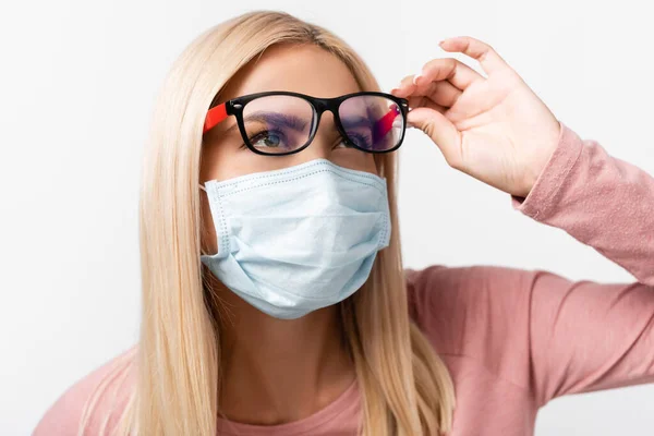 Mujer rubia en máscara médica sosteniendo gafas mientras mira hacia otro lado aislado en gris - foto de stock