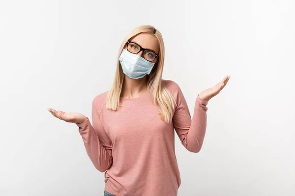 Blonde woman in medical mask and eyeglasses showing confused sign isolated on grey — Stock Photo