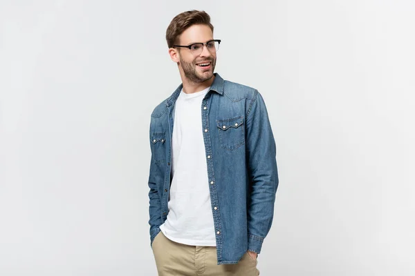 Hombre sonriente en gafas con las manos en los bolsillos de los pantalones mirando hacia otro lado aislado en gris - foto de stock