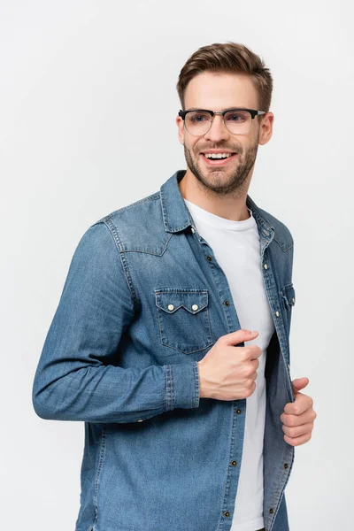 Молодий чоловік в окулярах посміхається, торкаючись джинсової сорочки ізольовано на сірому — стокове фото