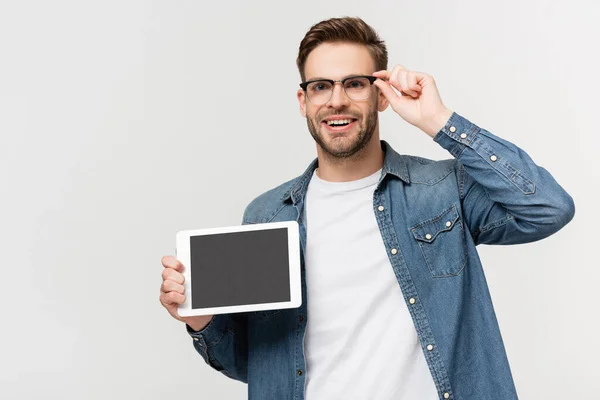 Uomo positivo toccare occhiali mentre tiene tablet digitale con schermo bianco isolato su grigio — Foto stock