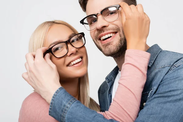Smiling couple looking at camera while touching eyeglasses of each other isolated on grey — Stock Photo