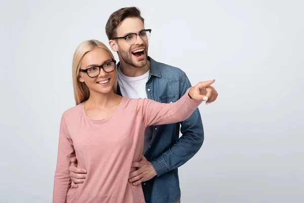 Cheerful man in eyeglasses embracing girlfriend pointing with finger isolated on grey — Stock Photo