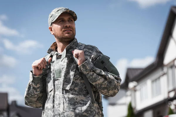 Erwachsener Soldat in Tarnkleidung schaut weg mit verschwommenen Häusern im Hintergrund — Stockfoto