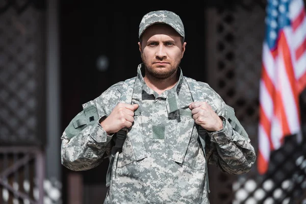 Ernsthafter Soldat in Uniform blickt in die Kamera mit verschwommenem Haus und Fahne im Hintergrund — Stockfoto