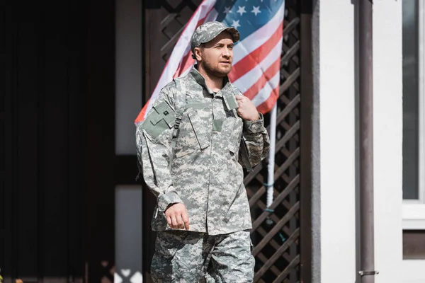 Militar confiado mirando hacia otro lado mientras está de pie cerca de la casa y la bandera americana - foto de stock