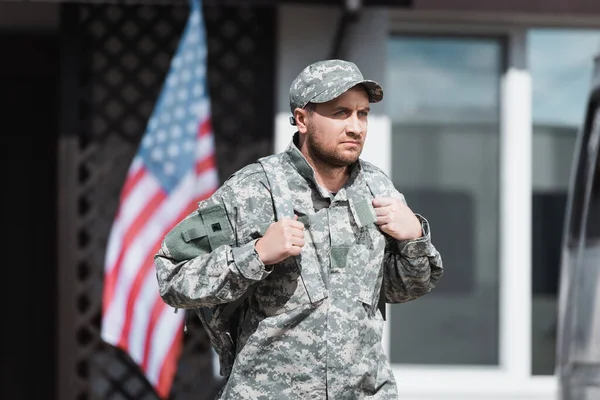 Hombre militar serio en camuflaje mirando hacia otro lado con la casa borrosa y la bandera de EE.UU. en el fondo - foto de stock