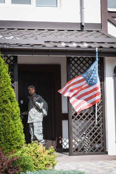 Un militar serio parado cerca de la puerta de la casa y mirando hacia otro lado cerca de los arbustos y la bandera americana - foto de stock