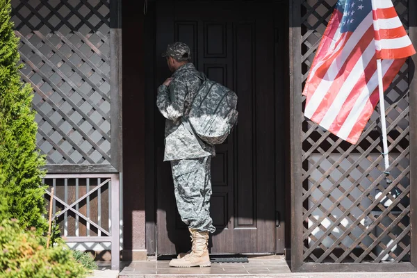 Back view of military man with backpack standing near house door and american flag — Stock Photo