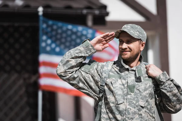 Militar sonriente en uniforme mirando hacia otro lado y saludando con la borrosa bandera de EE.UU. en el fondo - foto de stock