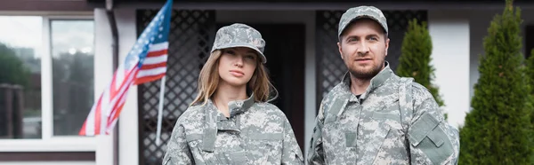 Military couple in uniforms standing together and looking at camera near house, banner — Stock Photo