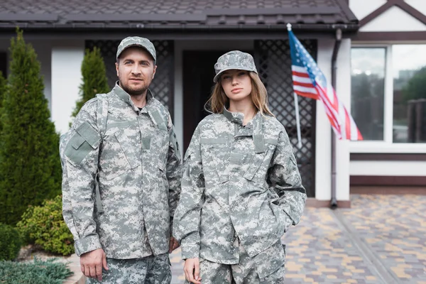 Pareja militar en uniformes de pie juntos y mirando a la cámara cerca de la casa - foto de stock