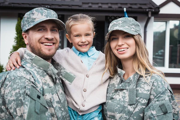 Feliz padre y madre en uniformes militares levantando a su hija y mirando a la cámara en un fondo borroso - foto de stock