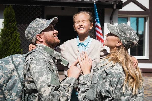 Happy daughter embracing father and mother in military uniforms near house with usa flag — Stock Photo