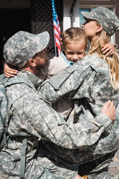 Розчарована дочка обіймає маму й батька у військовій формі біля дому з американським прапором. — стокове фото