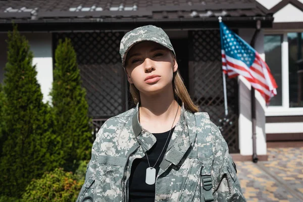 Femme militaire confiante avec collier regardant la caméra avec maison floue et drapeau américain sur fond — Photo de stock