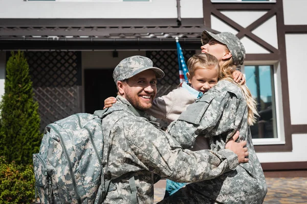 Smiling daughter hugging father and mother in military uniforms near house with american flag — Stock Photo