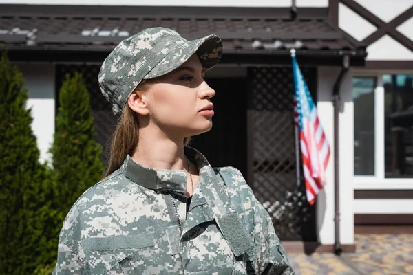 Perfil de mujer militar confiada en uniforme mirando hacia otro lado con la bandera americana borrosa y la casa en el fondo - foto de stock