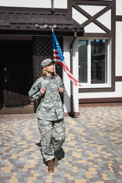 Ernste Soldatin mit Rucksack verlässt Haus mit amerikanischer Flagge im Hintergrund — Stockfoto