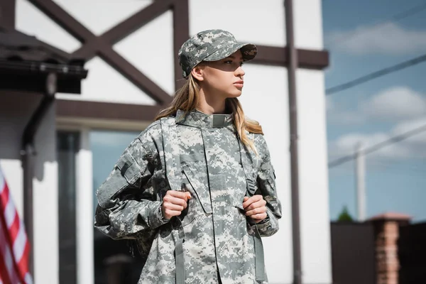 Confident military servicewoman with backpack looking away with blurred house on background — Stock Photo