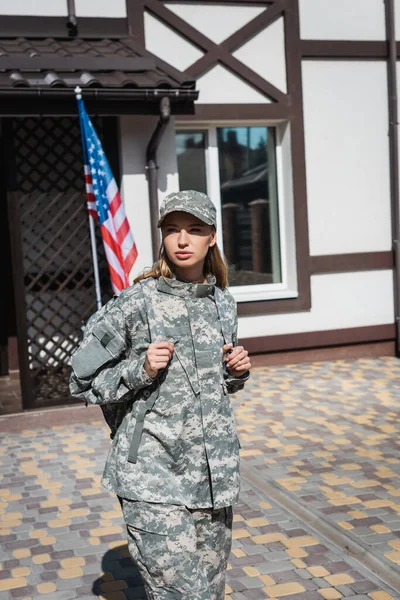 Serious military servicewoman leaving house and looking away with usa flag and house on background — Stock Photo