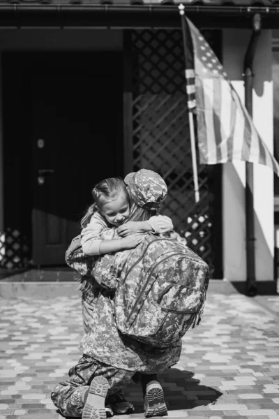 Military servicewoman embracing daughter, while sitting on knee near house and american flag, monochrome — Stock Photo