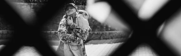 Happy mother in military uniform hugging daughter with blurred net on foreground, banner, monochrome — Stock Photo