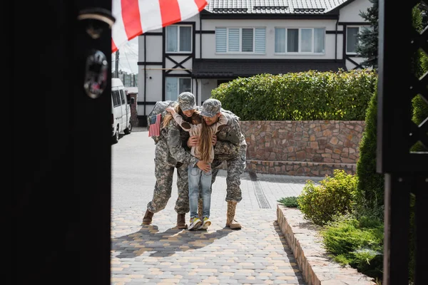 Tochter umarmt Mutter und Vater in Militäruniformen auf Straße nahe Haus im verschwommenen Vordergrund — Stockfoto