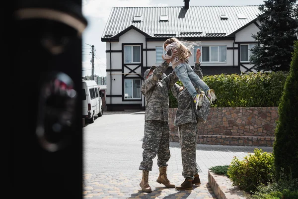Père et mère en uniforme militaire soulevant leur fille dans les airs, debout dans la rue au premier plan flou — Photo de stock