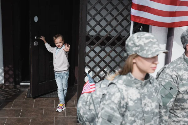 Smiling daughter with waving hand standing in doorway near american flag with blurred mother and father in military uniforms on foreground — Stock Photo
