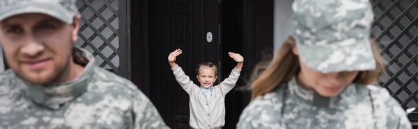 Happy girl with waving hands standing near house door with blurred man and woman in military uniforms on foreground, banner — Stock Photo