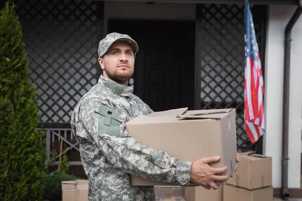 Military serviceman holding cardboard box, while looking away near house and american flag — Stock Photo