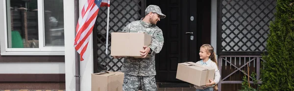 Père en uniforme militaire et fille tenant des boîtes en carton près de la maison avec drapeau américain, bannière — Photo de stock