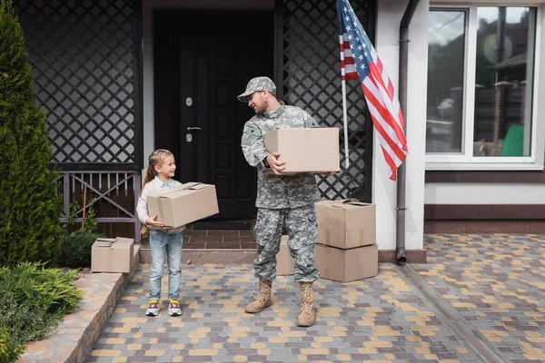 Father in military uniform and daughter holding cardboard boxes near house with american flag — Stock Photo