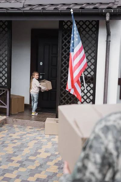 Menina sorridente com caixa de papelão em pé perto da porta da casa com pai desfocado em primeiro plano — Fotografia de Stock