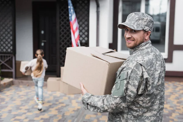 Feliz militar militar sosteniendo caja de cartón y mirando a la cámara con chica borrosa en el fondo - foto de stock
