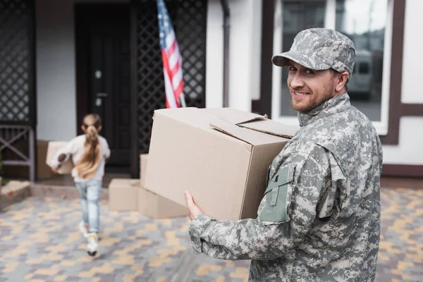 Heureux militaire tenant boîte en carton et regardant la caméra avec fille floue sur fond — Photo de stock