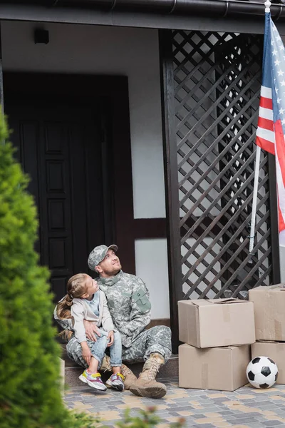 Father in military uniform hugging daughter, while sitting on threshold and looking at american flag — Stock Photo
